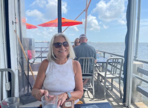 A woman with blonde hair and sunglasses smiles at a table by the water, with umbrellas and a sunny sky in the background.