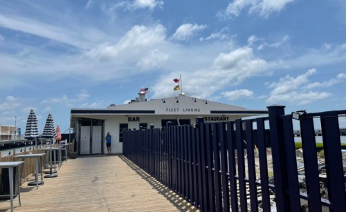 A waterfront restaurant with a bar, featuring flags and a wooden walkway under a blue sky with clouds.