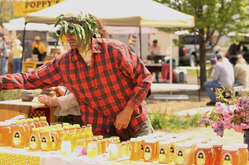 A man in a plaid shirt and leaf headpiece arranges jars of honey at an outdoor market.