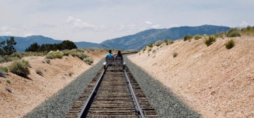 Two people ride a rail cart along a deserted train track, surrounded by mountains and greenery under a blue sky.