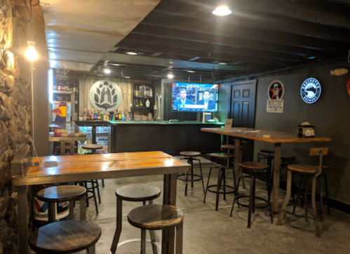 A cozy bar interior with wooden tables, stools, and a TV showing a sports game. Stone walls and dim lighting create a warm atmosphere.