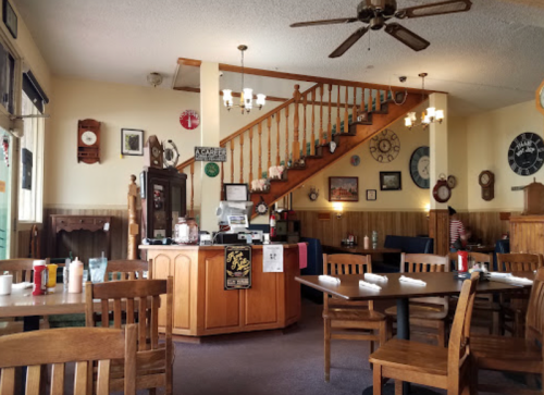 Cozy restaurant interior with wooden tables, a staircase, and various clocks on the walls. A counter is in the foreground.