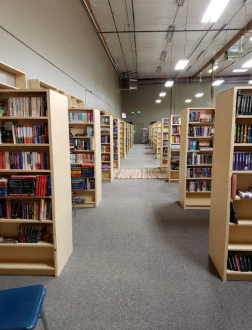 A spacious library aisle lined with tall bookshelves filled with books, leading to a distant area with more shelves.