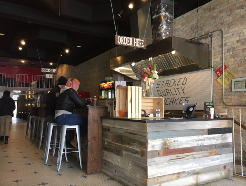 A cozy café interior with a wooden counter, patrons seated, and a sign that reads "Order Here." Flowers decorate the space.
