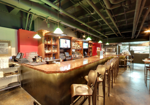 A modern bar with a polished countertop, wooden shelves, and seating, featuring TVs and a well-stocked liquor display.