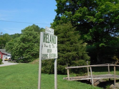 Sign reading "Welcome to Ireland, Home of the Irish Spring Festival," surrounded by greenery and a wooden bridge.