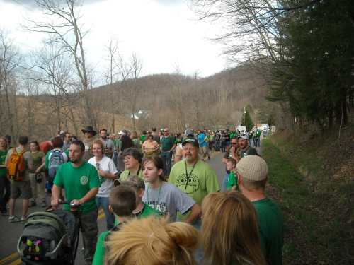 A crowd of people wearing green shirts walks along a rural road, surrounded by trees and hills.
