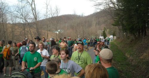 A large crowd of people in green clothing walking along a rural road, surrounded by trees and hills.
