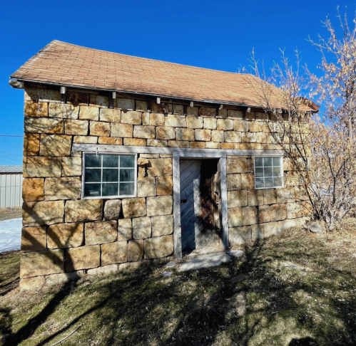 A rustic stone building with a sloped roof, featuring two windows and a wooden door, surrounded by sparse vegetation.