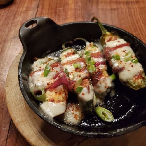 A cast iron skillet with three stuffed jalapeños wrapped in bacon, topped with melted cheese and green onions.