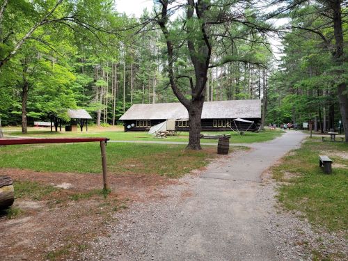 A path leads through a wooded area to a rustic building surrounded by trees and picnic tables.