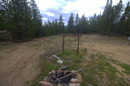 Sign marking the end of a motorized route, surrounded by trees and a fire pit on a dirt path.