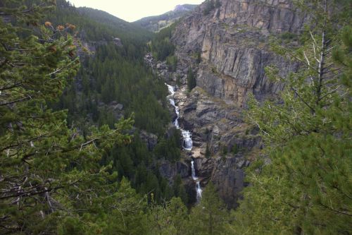 A scenic view of a waterfall cascading through rocky terrain, surrounded by lush green trees and mountains.
