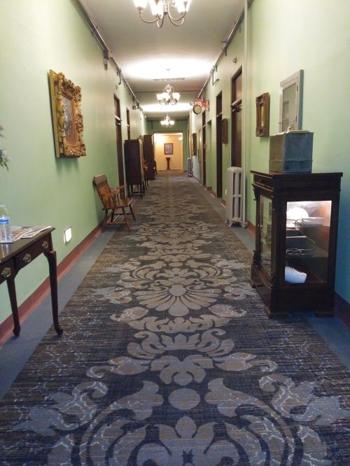 A long hallway with green walls, patterned carpet, and vintage furniture, illuminated by hanging lights.