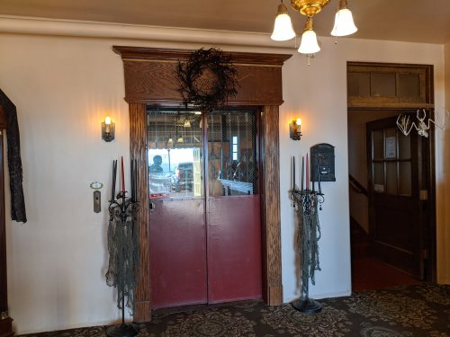 Double doors with a wreath, flanked by candle holders, leading to a rustic interior with warm lighting and wooden accents.