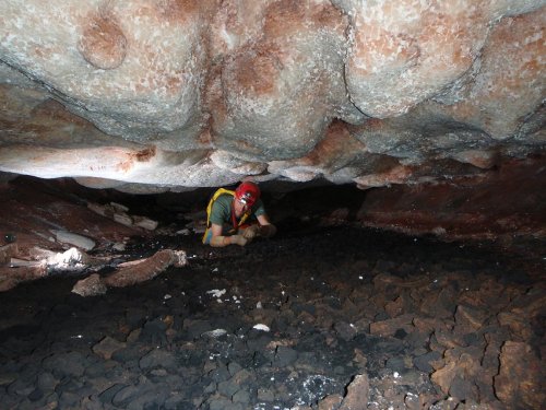 A person in a helmet crawls through a narrow cave passage with rocky ground and textured ceiling above.