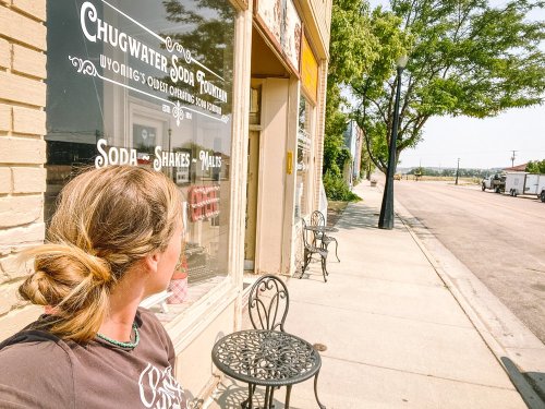 A person with a bun hairstyle sits outside a vintage soda shop, looking down a sunny street with empty chairs nearby.