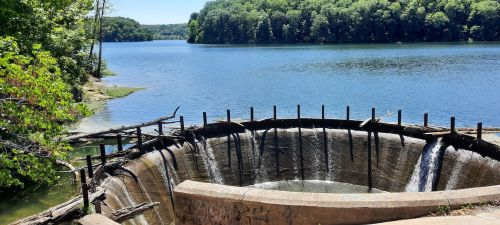 A serene lake scene featuring a circular dam with water cascading over its edge, surrounded by lush greenery.