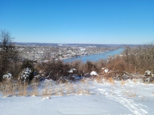 A snowy landscape overlooking a river, with a clear blue sky and distant hills in the background.