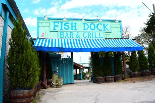 A colorful sign for "Fish Dock Bar & Grill" with a blue and white striped awning and greenery around the entrance.