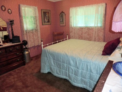 Cozy bedroom with a bed, wooden dresser, lace curtains, and pink wallpaper. Soft lighting creates a warm atmosphere.