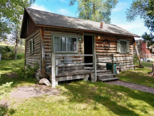 A rustic log cabin with a porch, surrounded by greenery and trees under a blue sky.