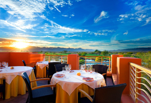 A scenic outdoor dining area with tables set for dinner, overlooking a sunset and mountains in the background.