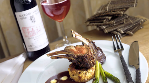 A gourmet plate featuring rack of lamb, asparagus, and a wine glass, with a bottle of red wine in the background.