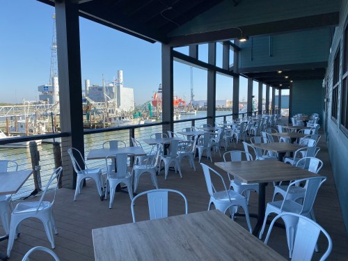 Outdoor dining area with empty tables and chairs overlooking a harbor and industrial scenery on a clear day.