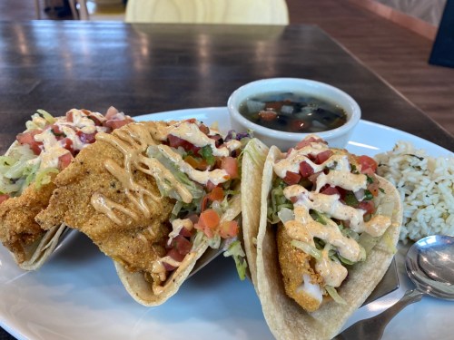 Three fish tacos filled with lettuce, tomatoes, and sauce, served with a side of black bean soup and rice.