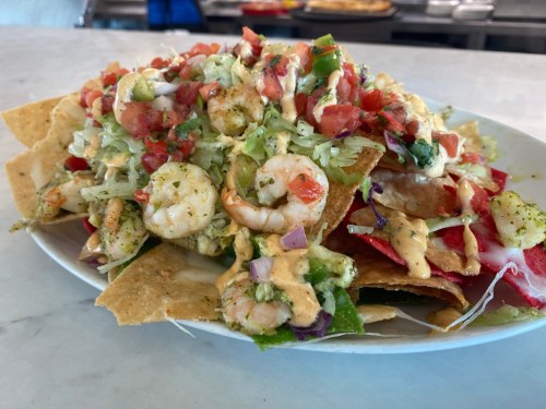 A plate of nachos topped with shrimp, lettuce, tomatoes, and a creamy sauce, served in a casual dining setting.