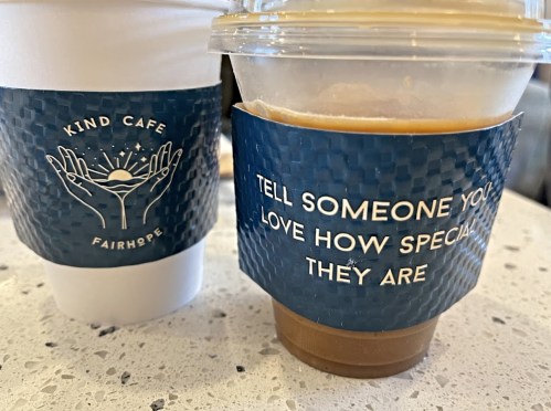 Two coffee cups on a table: one paper cup and one plastic, both featuring messages about kindness and love.