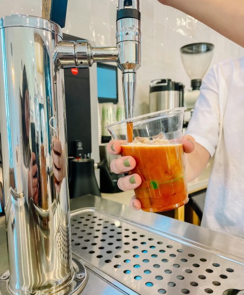A person pours a drink from a tap into a clear cup, with reflections visible on the shiny surface.