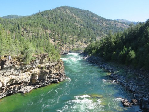 A scenic river flows through a lush green valley, surrounded by mountains and rocky cliffs under a clear blue sky.
