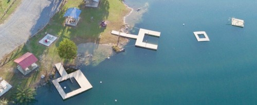 Aerial view of a lake with wooden docks and grassy areas, surrounded by trees and a few small structures.