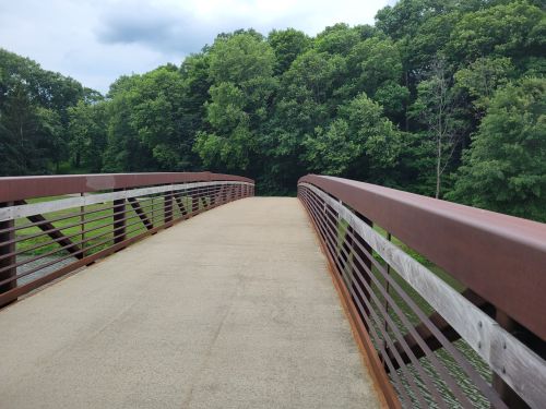 A metal bridge with wooden railings spans a calm area, surrounded by lush green trees under a cloudy sky.