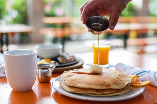 A hand drizzles syrup over a stack of pancakes with butter, alongside a cup of coffee and a glass of orange juice.