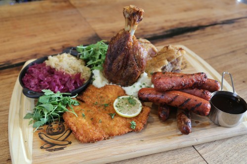 A wooden platter featuring a variety of meats, including a drumstick, sausages, and schnitzel, with sides of sauerkraut and greens.