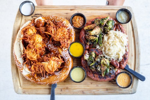 A wooden platter with two large pretzels topped with crispy potatoes and a variety of dipping sauces, alongside grilled vegetables.