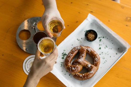 Two hands raise glasses of beer over a large pretzel on a wooden table, with dipping sauces on the side.