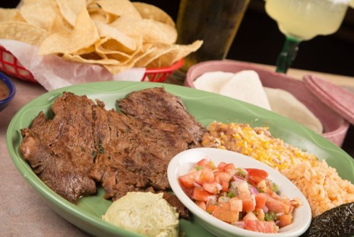 A plate of grilled meat with rice, beans, guacamole, pico de gallo, and tortillas, accompanied by chips and a drink.