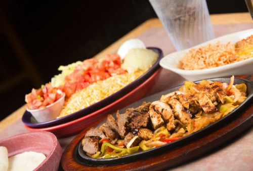 A sizzling platter of grilled meats with peppers, served alongside rice, salad, and tortillas.