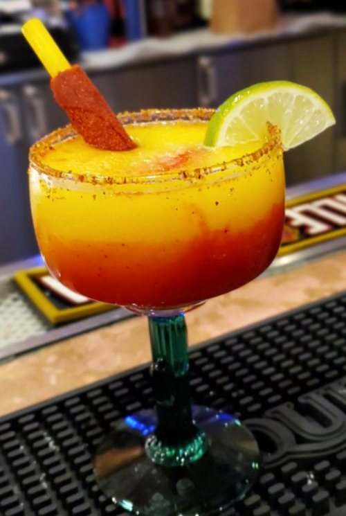 A colorful layered cocktail in a rimmed glass, garnished with a lime wedge and a stick, on a bar counter.