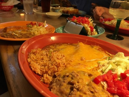 A colorful plate of enchiladas topped with cheese, served with rice, beans, and a side of salad on a restaurant table.
