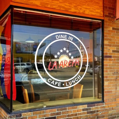 Window of "La Arepa" cafe featuring a circular logo with text "Dine In Cafe Lunch" on a brick wall.