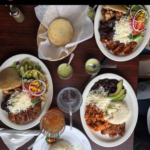 Three plates of colorful Mexican food with rice, beans, grilled chicken, avocado, and fresh vegetables on a wooden table.