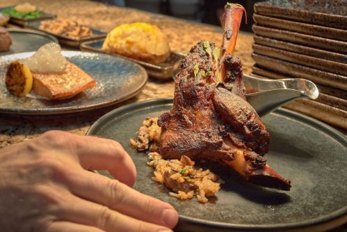 A beautifully plated dish featuring a large, roasted meat shank on a black plate, accompanied by rice and garnishes.
