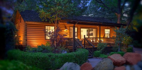 A cozy log cabin illuminated at dusk, surrounded by lush greenery and colorful rocks in the garden.