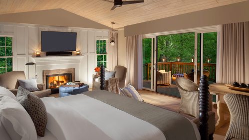 Cozy bedroom with a fireplace, large windows, and a deck overlooking greenery, featuring a bed and comfortable seating.