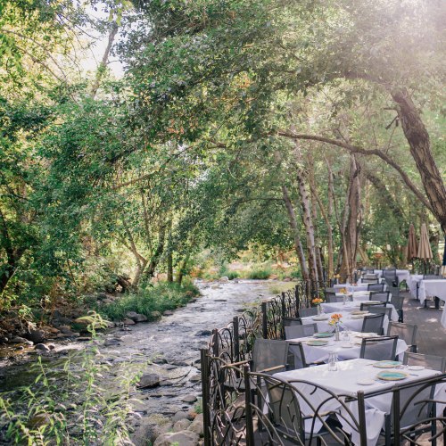 A serene outdoor dining area by a stream, surrounded by lush greenery and sunlight filtering through the trees.